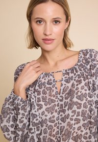 Lett og gjennomsiktig bluse i grå og brun leopardmønster. Har en rynket halslinning, nøkkelhullsåpning og vide ermer.