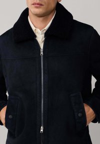 Marineblauwe faux-suède jas met een zwarte schapenvachtkraag, rits aan de voorkant en zijzakken met ronde zwarte knopen. Zachte textuur, getailleerde pasvorm.