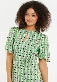 Vihreä kuvioitu mekko lyhyillä hihoilla, jossa on avainaukkoyksityiskohta ja vyötärönauha. Kangas on sileä ja siinä on geometrinen kuvio.