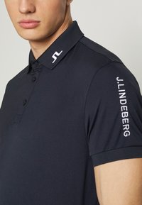 Camisa polo negra con cuello, que presenta una tapeta de tres botones, textura suave y un logo blanco en el cuello y la manga.