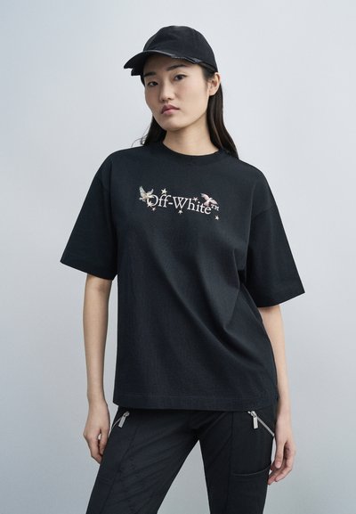 Joven mujer con camiseta negra Off-White con diseños de pájaros, pantalones negros con cremallera y gorra de béisbol negra, de pie frente a un fondo gris.