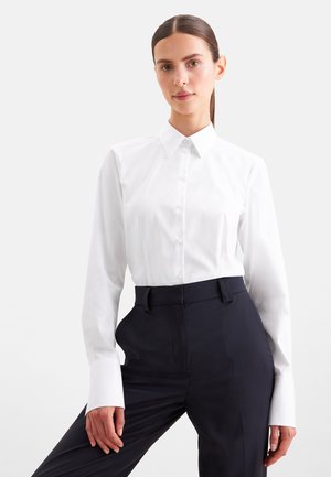 Kvinde med mørkt hår iført en tætsiddende hvid skjorte med knapper og højhælede sorte bukser, der står mod en ensfarvet lys baggrund.