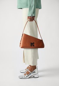 Brown triangular handbag with black logo, paired with white tailored pants and metallic silver heeled shoes, showcasing sleek design and textures.