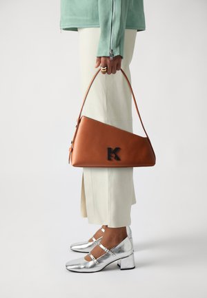 Hnědá trojúhelníková kabelka s černým logem, spárovaná s bílými tailored kalhotami a metalickými stříbrnými podpatky, které předvádějí elegantní design a textury.