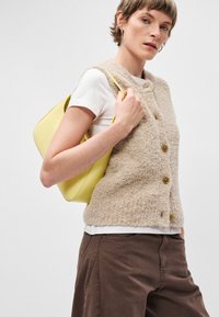 Bežna volna brezrokavnik z teksturirano tkanino, tremi gumbi in krepkim srajcem spodaj. Model nosi gladko, rumeno usnjeno torbico. Rjave hlače.