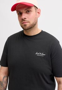 Άνδρας με ελαφριά γενειάδα, φοράει μπεζ και κόκκινο καπέλο, μαύρο t-shirt Jack & Jones και έχει τατουάζ στο αριστερό του αντιβράχιο, σε απλό λευκό φόντο.