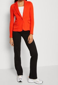 Blazer rosso su misura con due tasche frontali, abbinato a un top bianco e pantaloni a campana neri. Il modello indossa sneakers argentate con accenti arancioni.