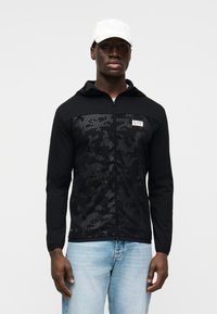 Μαύρο φούτερ με φερμουάρ, με υφή καμουφλάζ στο μπροστινό μέρος, μονόχρωμα μαύρα μανίκια και ανακλαστικό λογότυπο.