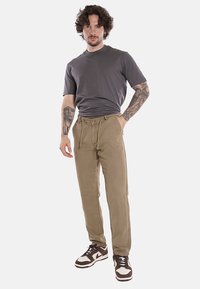 Maglietta grigia a girocollo, pantaloni khaki con cintura regolabile, e sneakers scure, in piedi su uno sfondo neutro.