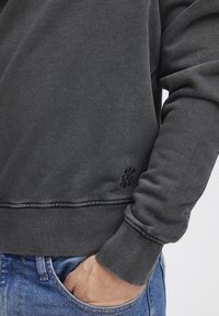 Sweatshirt gris avec un ourlet et des poignets côtelés, tissu texturé et un petit logo brodé près de la taille. Porté avec un jean bleu.