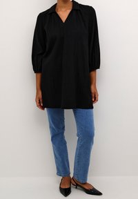 Personne portant une tunique noire à manches longues, un jean droit bleu et des escarpins noirs à bout pointu, se tenant devant un fond uni.