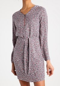 Robe fleurie en tissu léger, avec un décolleté en V, des manches longues, une ceinture à nouer à la taille et un motif multicolore avec des accents rouges et bleus.