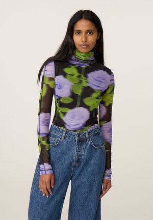 Vrouw met lang zwart haar die een doorschijnend zwart coltop draagt met grote paarse bloemen en groene bladeren, gecombineerd met een blauwe spijkerbroek.