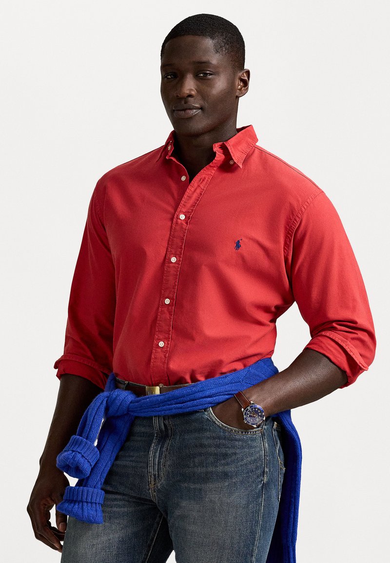 Camisa vermelha de colarinho com mangas longas, apresentando um logotipo azul. Suéter azul de malha amarrado à cintura, usado sobre jeans de ganga.