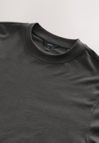 T-shirt in cotone grigio scuro con collo a giroclassico, maniche corte e una texture liscia. Presenta un'etichetta con "NEXT" e taglia "M".