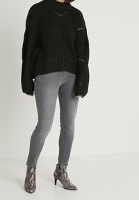 Pull en maille épaisse vert foncé avec un col montant, associé à un jean skinny gris et des bottines à imprimé serpent avec un bout pointu.