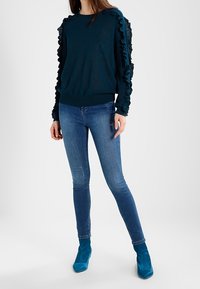 Türkiser Pullover mit Rüschensleeves, kombiniert mit eng anliegenden blauen Jeans und türkisen High Heels mit Knöchelschaft. Glatte Textur, lockere Passform.