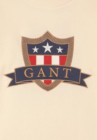 Bežová mikina se námornickým štítem, červenými a bílými pruhy, zlatými akcenty a nápisem "GANT" v tučném písmu.