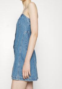 Femme portant une mini-robe en denim bleu sans manches avec de fines bretelles, debout de profil contre un fond blanc uni.