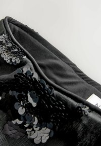 Sort stofpose med skinnende sorte palietter arrangeret i blomsterlignende mønstre, der viser detaljeret tekstur og sømning langs kanten mod en lys baggrund.