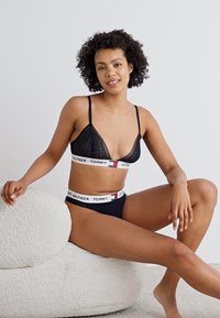 Zwarte kant bralette met dunne bandjes, gecombineerd met marineblauwe ondergoed met een witte band met het "TOMMY HILFIGER" logo in rode en blauwe accenten.