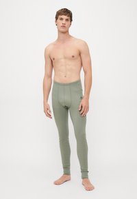 Uomo a torso nudo in piedi, scalzo e con addosso leggings lunghi aderenti di colore verde oliva, contro uno sfondo bianco semplice.