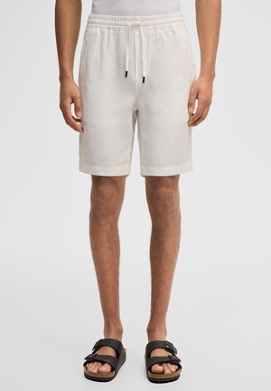 Man met witte shorts met trekkoord en zwarte sandalen met gesp, staand tegen een effen achtergrond.