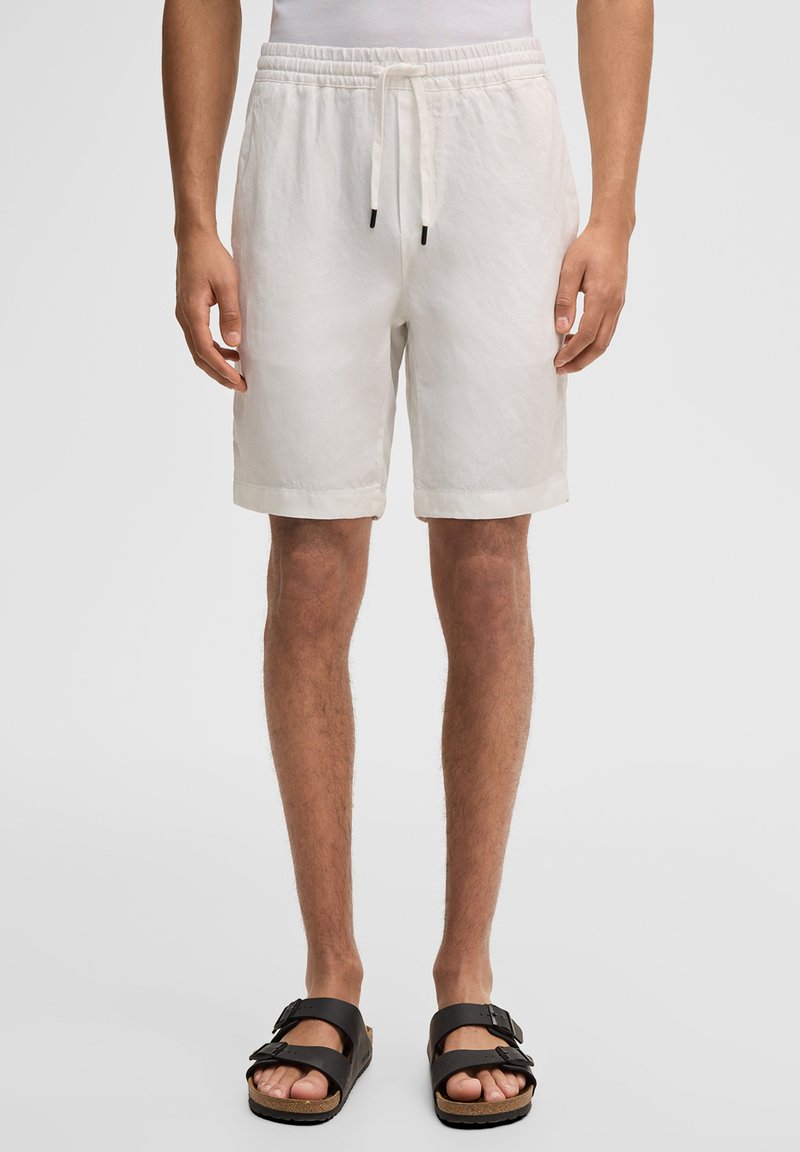 Homme portant un short blanc à cordon de serrage et des sandales noires à boucle, debout devant un fond uni.