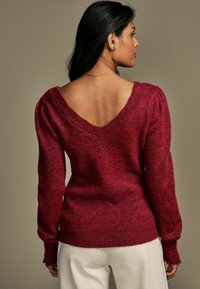 Maglione rosso lavorato a maglia con un ampio scollo a V, maniche a palloncino e orlo a costine. Texture morbida e colore ricco con leggere variazioni.