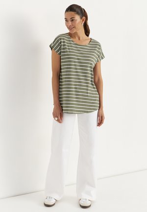 Kvinde med opsat hår iført en grøn- og hvidstribet t-shirt, hvide bukser med vide ben og hvide sneakers, stående og smilende.