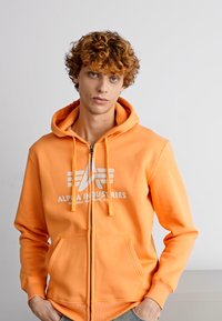 Narancssárga cipzáros kapucnis pulcsi, pamutból készült, elöl grafikai logóval, húzózsinóros kapucnival és oldalsó zsebekkel. Simaság és testhez álló fazon jellemzi.