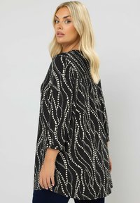 Svart tunika med vitt abstrakt swirl-mönster, 3/4-ärmar, avslappnad passform, tillverkad av lättviktsmaterial. Kännetecknas av sin flödande design och textur.