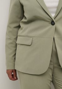 Světl zelený přiléhavý blazer s jediným černým knoflíkem, nakládanou kapsou a hladkým povrchem, doplněný o odpovídající kalhoty.