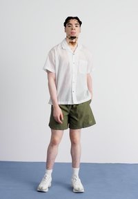 Camisa branca de manga curta com riscas verticais e bolso, combinada com calções verde-oliva. Meias brancas e sapatilhas brancas completam o conjunto.
