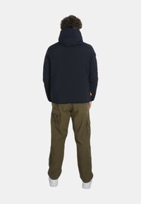Giacca imbottita navy con cappuccio, abbinata a pantaloni cargo oliva con tasche e sneakers bianche, vista da dietro.