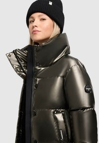 Metallic oliven grøn pufferjakke med høj krave, trykknapper og sidelommer. Båret med en sort ribbet strikhue.