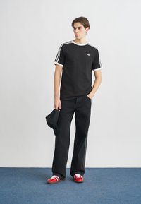 Čierne bavlnené tričko s bielymi pruhmi na rukávoch, doplnené voľnými čiernymi džínsami a červenými teniskami. Model drží čiernu šiltovku.