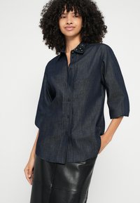 Tmavá denimová košeľa s predným zapínaním na gombíky, trojštvrťovými rukávmi a ozdobnými detailmi na golieri. Hladká textúra a pohodlný strih.