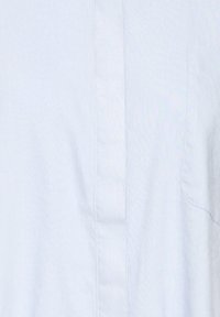 Gros plan d'une chemise bleu clair montrant la patte de boutonnage avant et la poche poitrine avec une texture subtile du tissu.