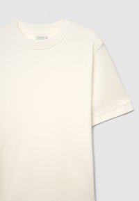 Schlichtes, kurzärmeliges, cremefarbenes T-Shirt mit Rundhalsausschnitt auf einem Bügel vor einem hellgrauen Hintergrund.