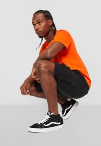 Baskets montantes noires avec des accents blancs, associées à un short cargo noir et un t-shirt orange, sur un fond gris uni.