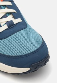 Blue sneaker with mesh and suede materials. Features cream laces and a textured sole. Upper has varying shades of blue and stitching details.