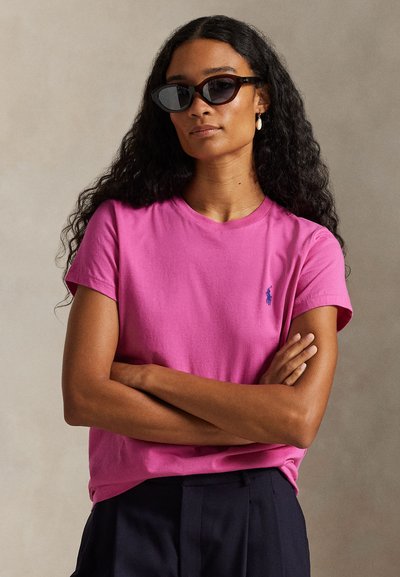 Maglietta rosa a maniche corte in cotone con scollo tondo e piccolo logo blu. Modello con occhiali da sole neri e pantaloni scuri, con le braccia incrociate.