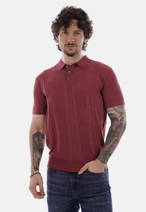 Mann mit lockigem Haar trägt ein kurzärmeliges, weinrotes Polohemd und blaue Jeans, zeigt detaillierte Tätowierungen an beiden Unterarmen, steht vor weißem Hintergrund.