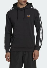 Svart hoodie med dragsko, känguruficka och vita tre-stripsdetaljer på ärmarna. Har en liten orange logotyp på bröstet.