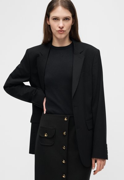 Γυναίκα με μαύρο σακάκι, μαύρο κοντομάνικο ριγέ τοπ και μαύρη φούστα με χρυσά κουμπιά και τσέπη, ποζάροντας με το ένα χέρι στη μέση μπροστά σε λευκό φόντο.