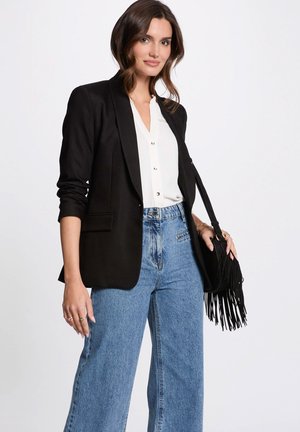 Frau, die einen schwarzen Blazer, eine weiße Bluse, hoch taillierte blaue Jeans trägt und eine schwarze Schultertasche mit Fransen bei sich hat.