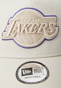 Justerbar beige kasket med et broderet "Los Angeles Lakers" logo i creme og lilla. Har et New Era mærke på skyggen.