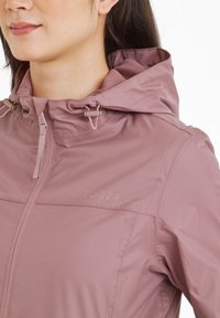 Lightweight, hooded jacket in soft mauve featuring a zip front, adjustable drawstrings, and textured interior lining with a logo.