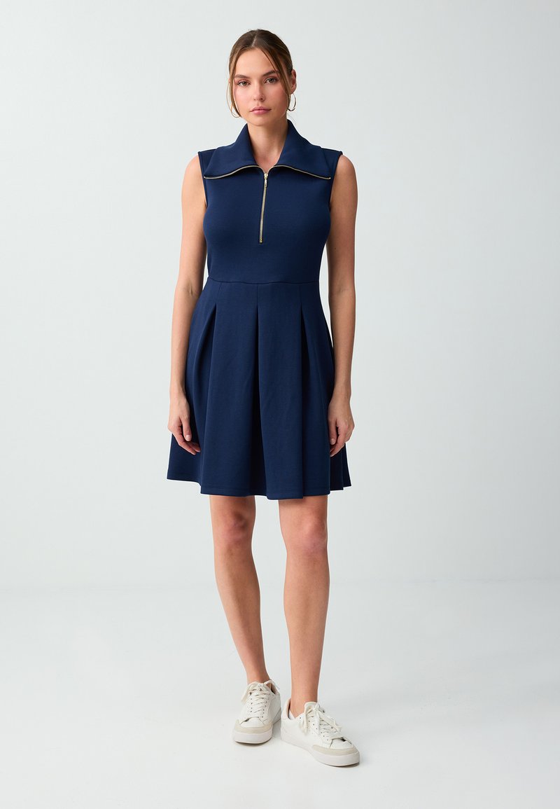 Femme debout portant une robe bleu marine sans manches avec une fermeture éclair à l'avant et des baskets blanches sur un fond clair uni.